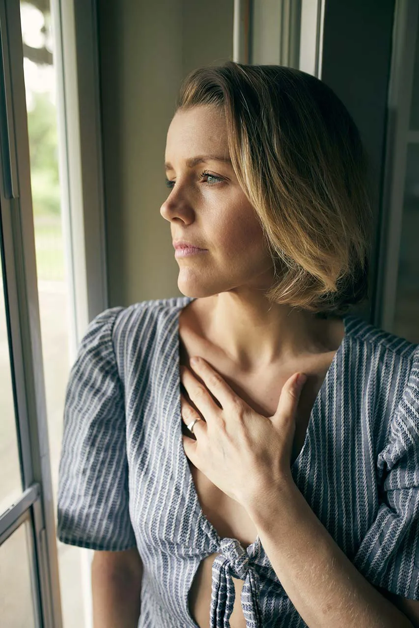 Woman standing at window with hand on heart - feeling overwhelmed